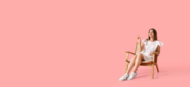 テキストのためのスペースを持つピンクの背景に何かを指している木製の肘掛け椅子のかなり若い女性 - 写真・画像