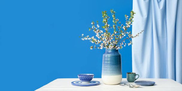 Florero con ramas de árbol florecientes y platos sobre mesa blanca sobre fondo azul. Banner para el diseño - Foto, imagen