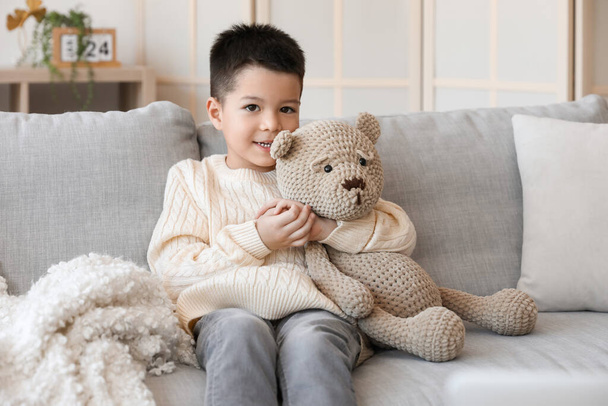 Aranyos kisfiú kötött pulóverben játékmacival otthon ül a kanapén. - Fotó, kép