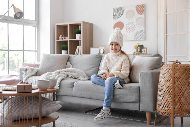Roztomilá holčička v pleteném klobouku a svetru sedí na pohovce doma - Fotografie, Obrázek