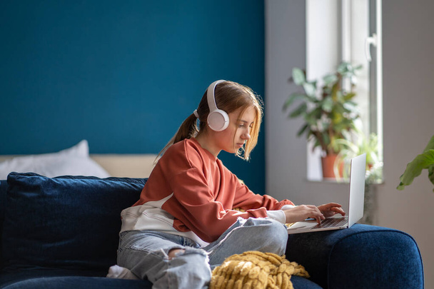 Фокусована дівчина-підліток в навушниках, що друкують текст на ноутбуці, розслаблена, сидить на дивані у вітальні. Студент школи-підлітка Pensive, який вивчає онлайн, дистанційне навчання, використовуючи комп'ютер вдома
. - Фото, зображення