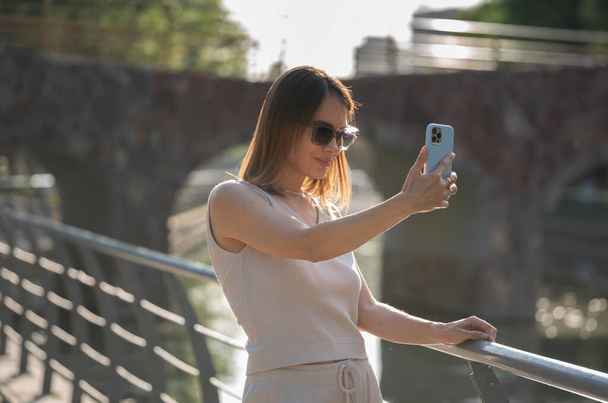 Φωτισμένο καλοκαιρινό πορτρέτο μιας νεαρής μελαχρινής γυναίκας με γυαλιά ηλίου. Μια γυναίκα στέκεται στη γέφυρα και βγάζει μια selfie σε ένα smartphone. Υψηλής ποιότητας φωτογραφία - Φωτογραφία, εικόνα