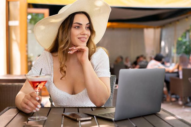 Young attractive woman wear dress and a straw hat drinks cocktails and works freelancer while sitting in cafe or on hotel terrace. Copy space - Photo, Image