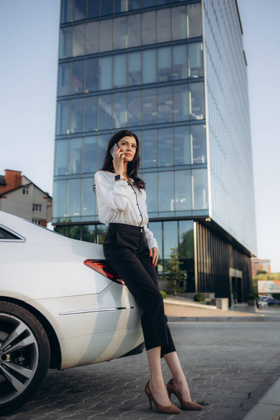 Mujer de negocios utiliza Smartphone mientras se apoya en su coche de primera clase. Gran ciudad con rascacielos en segundo plano
 - Foto, imagen