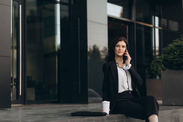 Attractive young businesswoman in white suit talking phone standing near business centre. - Photo, Image