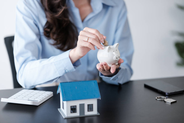 Close up Asian womens hand saving a coin into piggy bank with wooden house model on the table for business, finance, saving money and property investment concept. High quality photo - 写真・画像