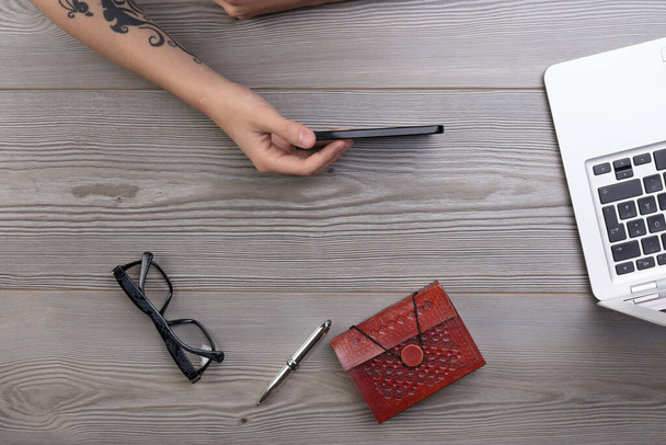 Bovenaanzicht van een vrouw met een smartphone op een houten bureau, laptop in de buurt. Bril, notitieboekje en pen ook aanwezig. Mengsel van modern en traditioneel lezen / schrijven - Foto, afbeelding