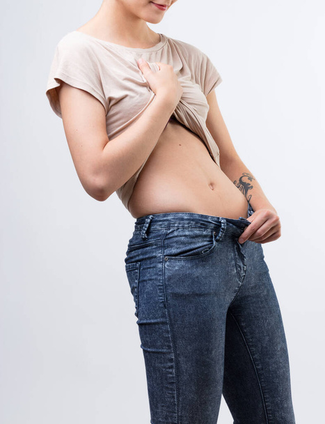 Młoda kobieta sprawdza swój idealny płaski żołądek. Nierozpoznawalna kobieta w obcisłych dżinsach, podnosi koszulę odsłaniając piękny brzuch. Siłownia, ćwiczenia, zdrowa dieta... Ona zawsze czuje się nieodpowiednia, pomimo jej  - Zdjęcie, obraz