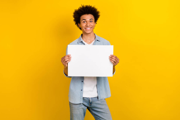 Portrait of toothy beaming nice guy wear jeans jacket holding white placard announce sale empty space isolated on yellow color background. - Foto, afbeelding