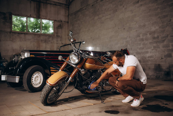 Ένας άνθρωπος σκουπίζει τη μηχανή μιας ρετρό μοτοσικλέτας. Retro συλλέκτη αυτοκινήτων και μοτοσικλετών. - Φωτογραφία, εικόνα
