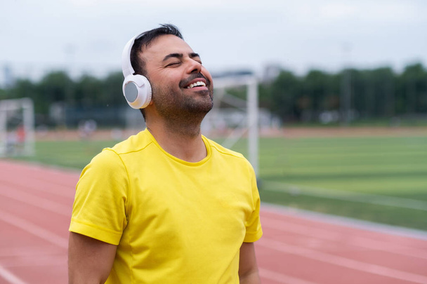 Usmívající se muž poslech oblíbené motivační píseň přes bezdrátové sluchátka školení na venkovním městském sportovním stadionu sportovec těší cvičení s hudbou - Fotografie, Obrázek