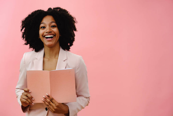 Portret van jonge zwarte vrouw met notebook, lachend en lachend, tegen roze studio achtergrond - Foto, afbeelding
