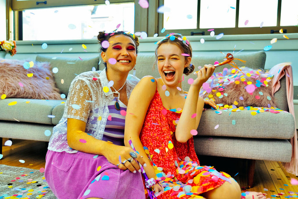 Two friends having party at home sitting on floor holding party blowers with confetti falling around them - Photo, Image