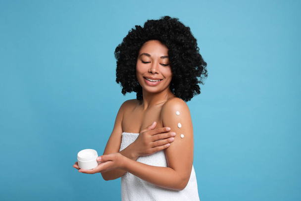 Young woman applying body cream onto shoulder on light blue background - Foto, afbeelding
