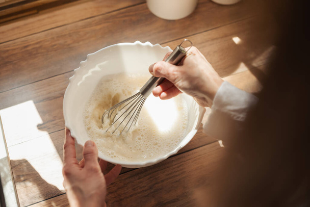 Girl whipping dough for baking bread. Close up of a wife cooking in kitchen, using whisk to stir different ingredients for liquid dough - Photo, Image