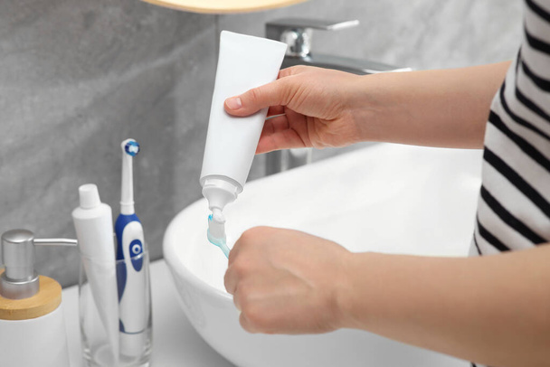 Mujer exprimiendo pasta de dientes del tubo al cepillo de dientes cerca del lavabo en el baño, primer plano - Foto, imagen