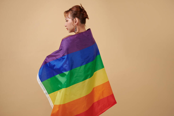 Serious transgender woman wrapped in rainbow flag standing against beige background - Fotó, kép