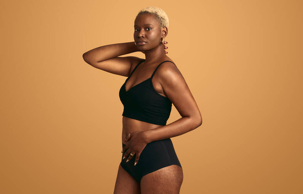 Self assured young African American female model wearing black underwear touching belly while standing against orange background - Fotografie, Obrázek