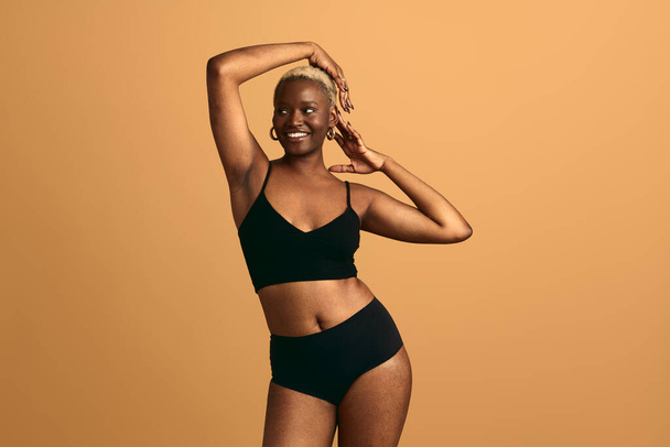 Cheerful young African American female model wearing black underwear with raised arms and short hair looking away against beige background in modern studio - Fotografie, Obrázek