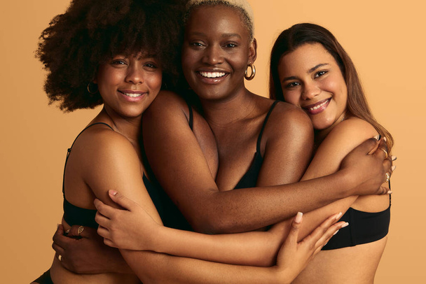 Cheerful young multiracial female friends in black underwear embracing and smiling while standing against yellow background together in studio - Foto, Bild