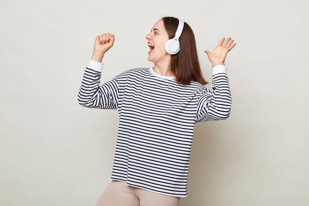 Attractive positive young woman wearing striped shirt listening to music in headphones isolated over gray background having fun celebrating raised arm as microphone singing. - Foto, afbeelding