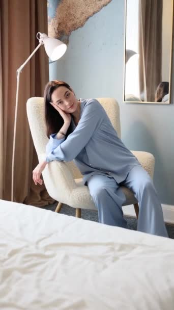 Jonge brunette vrouw in blauwe pyjama zit in een fauteuil in de slaapkamer tegen de achtergrond van thuis interieur. Verticale video. - Video