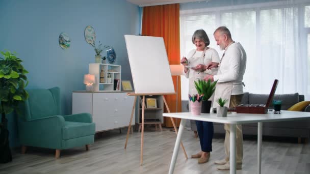 moderní důchodci rádi kreslí kreativní obrazy v penzi, stařec se ženou malují obraz štětcem a malbou doma - Záběry, video