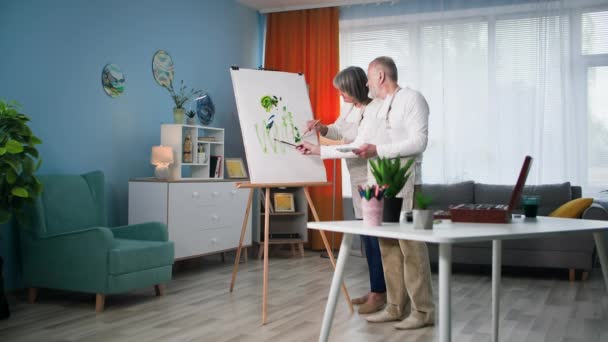 Развлечения на пенсии, пожилые мужчины и женщины художники рисуют современную картину кисточками и красками на холсте в уютной комнате - Кадры, видео