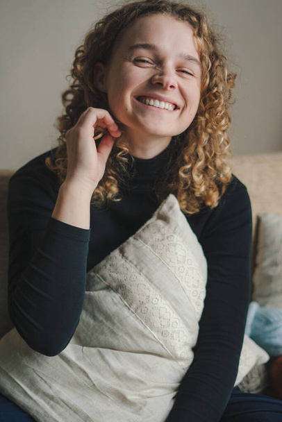 Pozitív fiatal nő göndör hajjal mosolyog és a kamerába néz, miközben otthon ül a kanapén a nappaliban - Fotó, kép