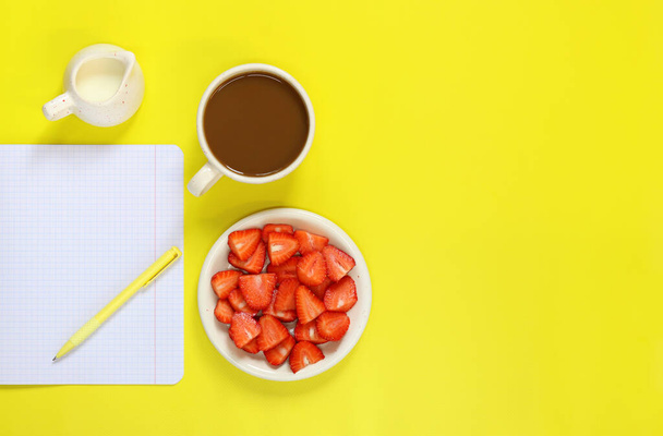 Cozy workspace. Mockup of an open notepad. Top view of a work desk, a notebook for notes, a cup of coffee, a milk jug, a pen and a plate with strawberries on a yellow background. Flat lay. - Photo, Image