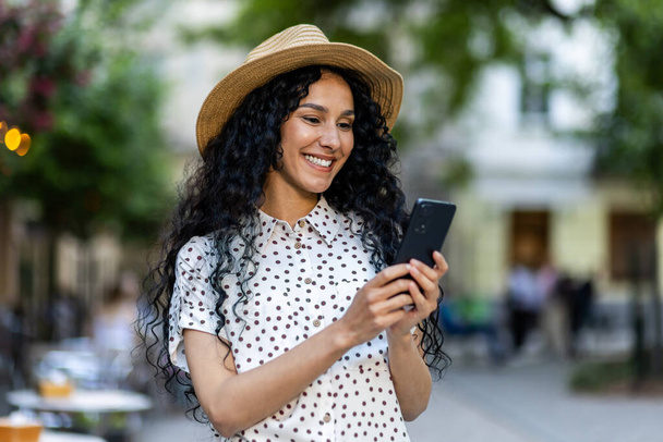 A beautiful young woman walks through the evening city in a hat, a smiling Latin American woman holds a smartphone in her hands. A tourist with curly hair types a message and browses online pages on - Fotoğraf, Görsel