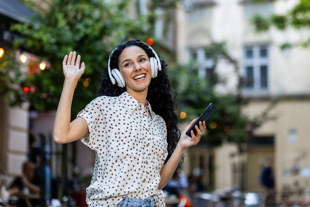 Beautiful young Latin American woman walks in the evening city on a trip, woman with headphones listens to music and dances and sings, uses an online radio application on her phone - Foto, immagini