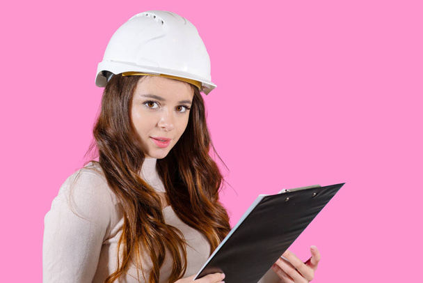 Μια νεαρή γυναίκα μηχανικός σε ένα κράνος κρατά το έργο της στα χέρια της στο ροζ φόντο. - Φωτογραφία, εικόνα
