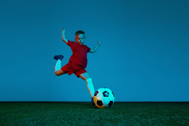 Male beginner soccer player in sports uniform in action, motion with ball over dark blue background in neon. Concept of action, sportive lifestyle, team game, health, energy, vitality and ad - Fotoğraf, Görsel