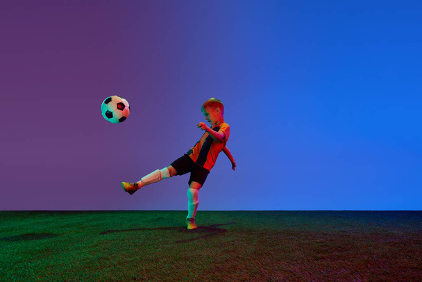 Male beginner soccer player in sports uniform in action, motion with ball over dark blue background in neon. Concept of action, sportive lifestyle, team game, health, energy, vitality and ad - Photo, Image