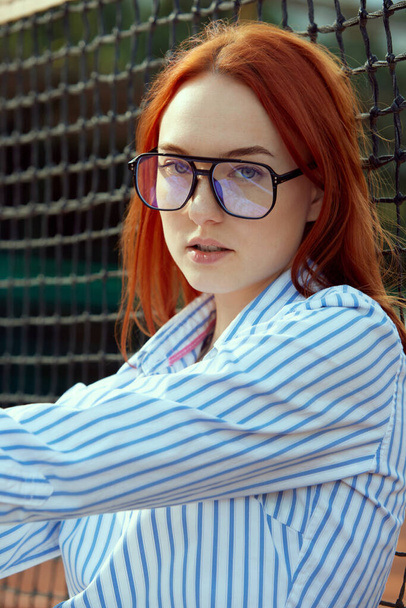 Close-up portrait of beautiful young redhead woman in stylish glasses and striped shirt posing near tennis net on sunny day outdoors. Casual fashion, sport, lifestyle, hobby, leisure time, ad concept - Photo, image