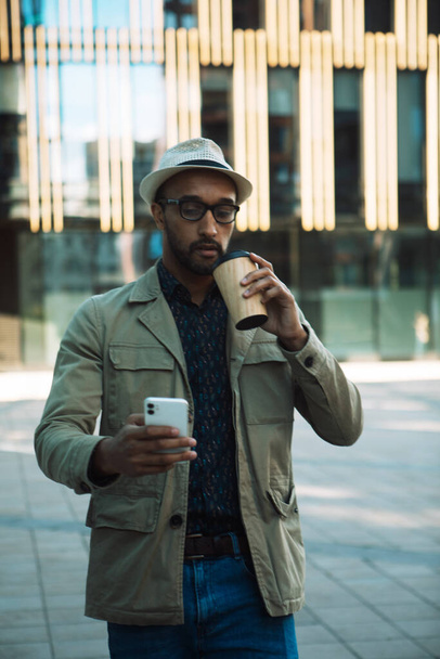 Young afro american man dressed casual holding a cup of coffee to go, smiling happy using smartphone at the city. Lifestyle and people concept. - Foto, Imagen