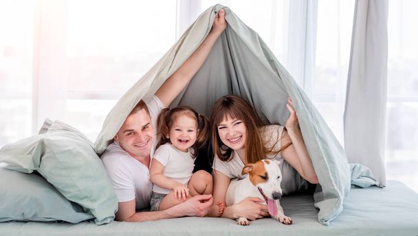 Посміхаючись сім'ї з собакою під ковдрою в ліжку. Прекрасні ранкові миті матері, батька і маленької дочки з домашнім улюбленцем під ковдрою
. - Фото, зображення