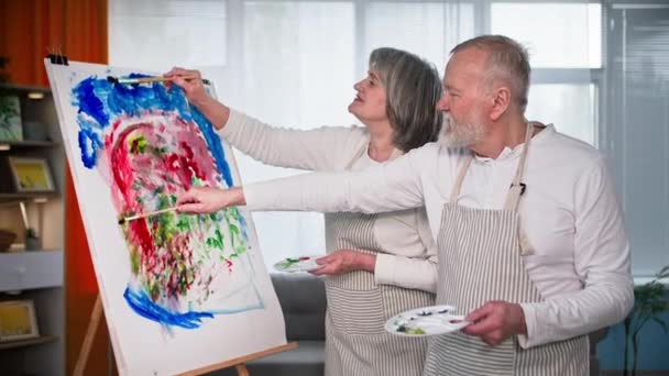 tehetséges nyugdíjasok, egy idős férfi és egy idős nő festeni kép ecsetek és festékek vászon a szobában - Felvétel, videó