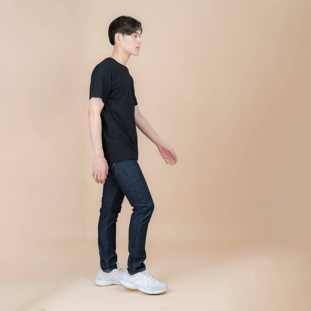 Male fashion model in black t-shirt and jeans standing in studio on beige background, trendy clothing style, copy space - Foto, Imagem