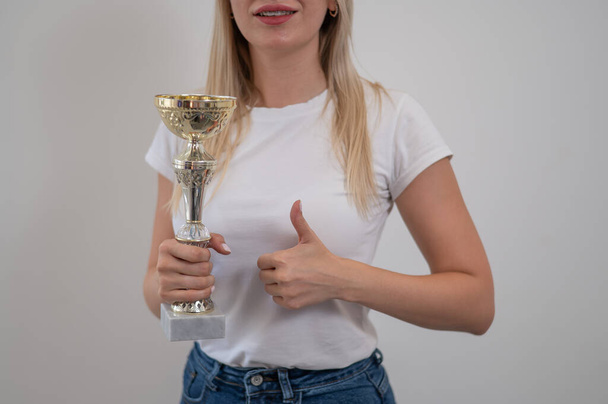 Blonde woman smiling and holding golden trophy cup and showing thumb up and against white wall - Fotografie, Obrázek