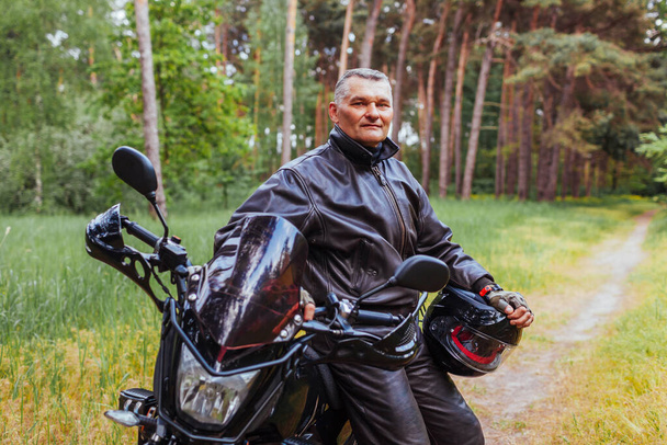 Elderly biker leaning on motobike in summer forest. Man in leather clothes standing with arms crossed enjoying nature. after ride - Photo, Image