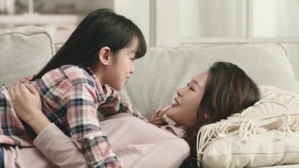 joven asiático madre y siete años de edad, hija acostado en sofá tener un conversación - Metraje, vídeo