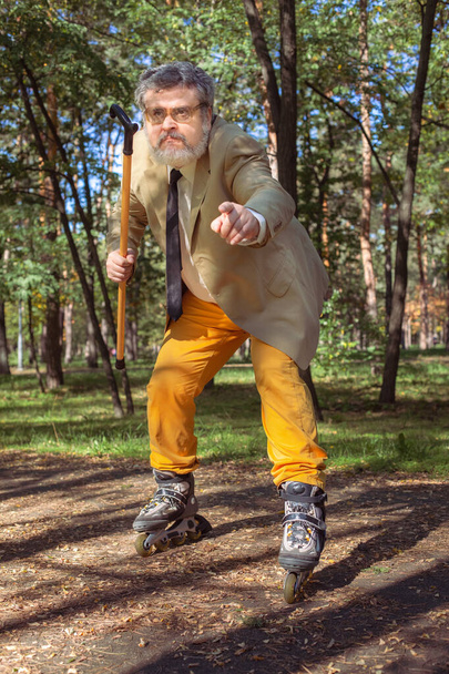 Zabawny staruszek jeździ na rolkach po parku. Dziadek z chodzącym kijem tańczy i robi miny. Dziadek z białą brodą. - Zdjęcie, obraz