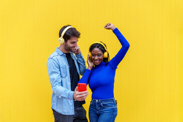 Em um tiro animado e colorido, um homem caucasiano e uma mulher africana groove à música, compartilhando risos e camaradagem com celulares e fones de ouvido. - Foto, Imagem
