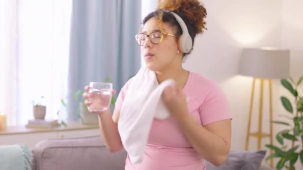 Słuchawki, ręcznik i kobieta pijące wodę po treningu w salonie swojego domu. fitness, nawodnienie i zmęczona młoda kobieta pocąca się na przerwie po ćwiczeniach z muzyką w domu - Materiał filmowy, wideo