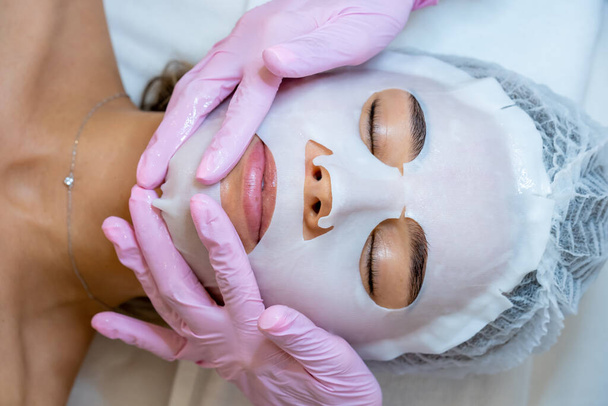 Unrecognizable professional doctor cosmetologist applying sheet mask on face of customer, woman in aesthetic clinic. - Φωτογραφία, εικόνα