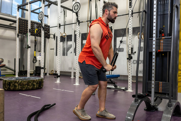 Bearded adult man working out at gym, doing cable rope pushdowns, biceps and triceps arm training workout. - Foto, Imagen