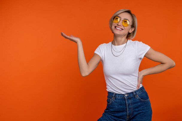 Charming smiling woman in white t shirt and jeans presenting advertisement area on her palm isolated over orange background. - Foto, afbeelding