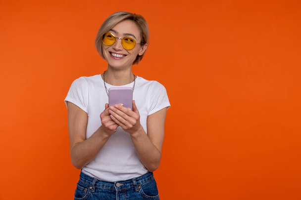 Positive woman wearing white T-shirt using mobile phone checking social networks isolated over orange background. - Foto, Imagem